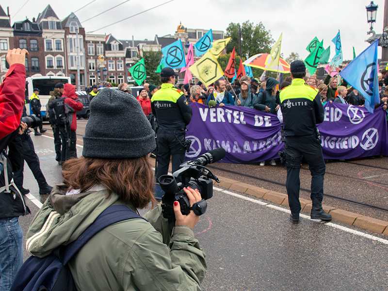 Fotograaf staat voor protesterende menigte om foto's te maken van 2 politieagenten die ervoor staan.
